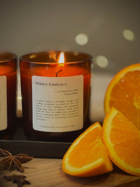 Candle labeled 'Winter Embrace' with orange slices and star anise on a wooden surface.